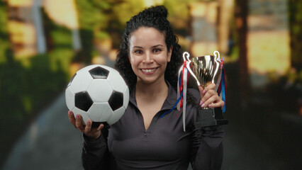 Obraz premium Woman holding soccer ball and trophy in sunny park smiling proudly after victory showcasing success outdoor sport achievement.