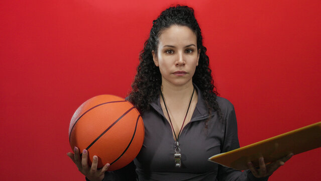 Hispanic woman coach holding a basketball and clipboard against a vibrant red wall, symbolizing sports leadership and strategy in an indoor setting.