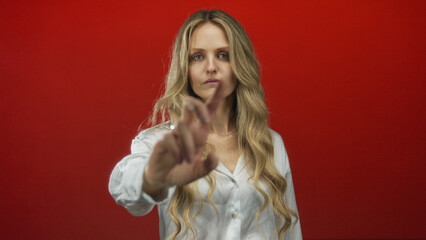 Woman with long blonde hair stands against a bold red background, extending her hand forward as if...