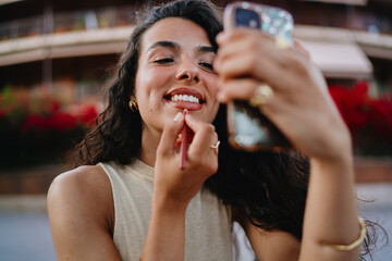 Smiling woman applies lip liner using smartphone as mirror, merging beauty routine with mobile technology for on-the-go self-care and digital convenience.