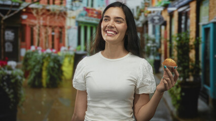 Woman smiling while holding muffin and looking upward wearing white shirt under bright sunlight on busy street; joy.