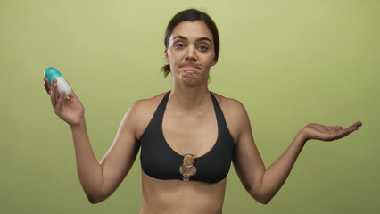 Woman holds roll on deodorant and shrugs with uncertain expression in olive green studio; personal...