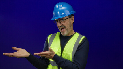 Man in hardhat and safety vest gesturing confidently against a blue background, depicting skilled professional engaging in a presentation or instruction setting.