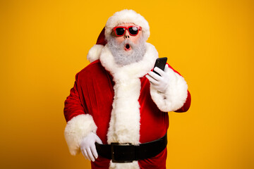 Funny Santa Claus in red costume using smartphone against vibrant yellow background expressing surprise with cool sunglasses