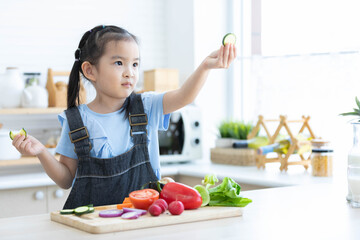 cute asian little child girl eating slice a cucumber with different vegetables in the kitchen
