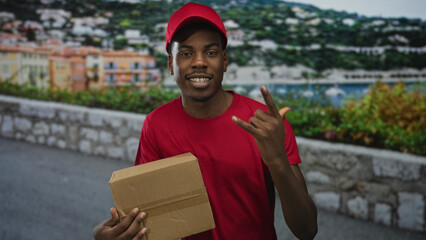 Fototapeta premium Man in red cap holds cardboard box and makes rockhorns gesture on street alongside stone wall and coastal buildings; confidence.
