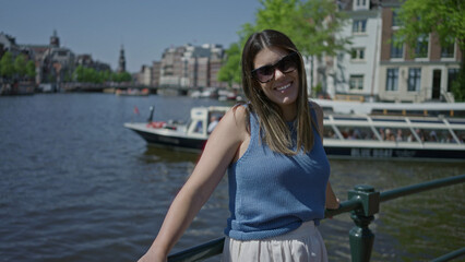 Fototapeta premium Woman enjoying sunlight by amsterdam canals with historic architecture in the background, wearing sunglasses, capturing a relaxed moment in the urban outdoors of the netherlands.