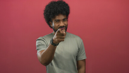 African american man pointing at camera with a confident smile, set against a solid red background,...