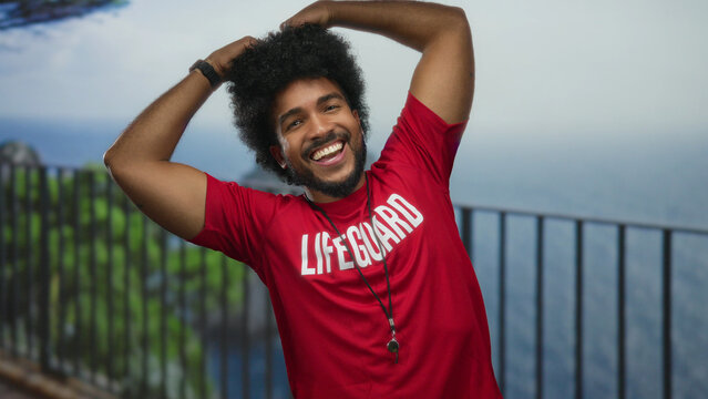 Lifeguard man smiling by the seaside promenade with heart gesture, wearing red uniform, against the scenic beach and sea outdoor backdrop.