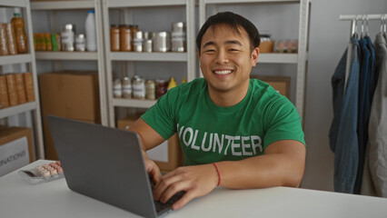 Young man volunteering in charity center room working on laptop wearing green shirt shelves in background