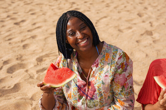 Happy young african woman eating fresh watermelon on the beach - Picnic, healthy food and vacation concept - Powered by Adobe