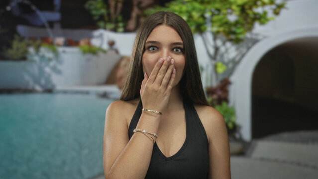 Girl covers her mouth with hand at resort poolside wearing black swimsuit top in bright sunlight; surprise.