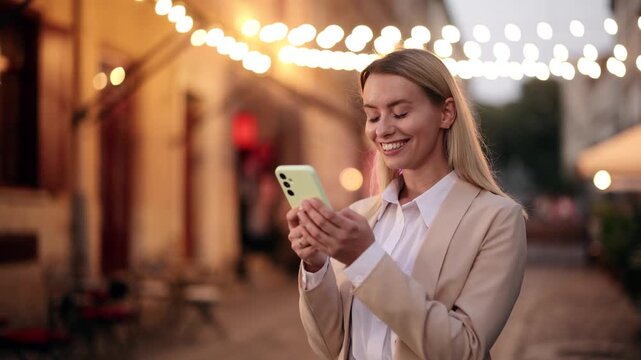Cheerful woman with smartphone under glowing string lights in evening city street, smiling and chatting online outdoors