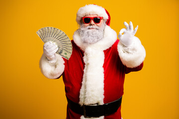 Santa Claus in a festive red hat holds a fan of money and gestures okay against a bright yellow background