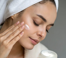 A woman holds cream in her hands and applies it to her face. The skin looks moisturized. The background is calm, neutral