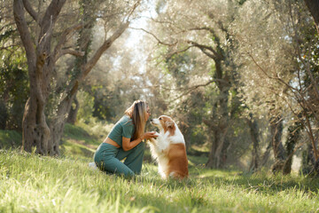 The woman presses her forehead to her Border Collie in quiet surroundings. Warm sunlight and tall trees frame their affectionate exchange.