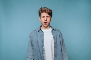 Fototapeta premium Young man in denim shirt showing surprise against blue backdrop, expressing shock in a moment of realization