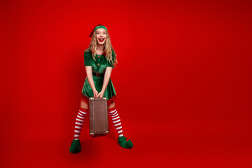 Cheerful holiday elf posing with vintage suitcase for Christmas party celebration on vibrant red background