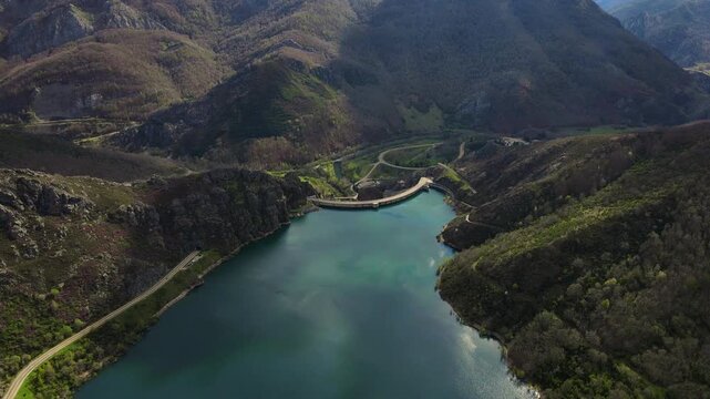 Aerial drone footage of the Riano reservoir and hydroelectric dam in Leon, Spain. The video captures turquoise waters surrounded by dramatic mountain peaks, the engineering structure of the dam, and