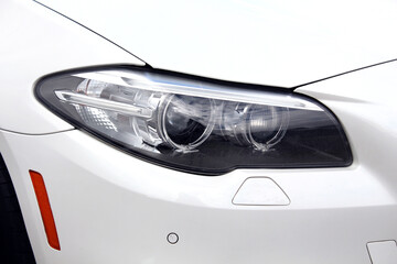 Modern headlights with dual projection lenses, chrome accents, and clear housing, integrated into white bumper with parking sensors. Modern headlights with headlight washers on white car.