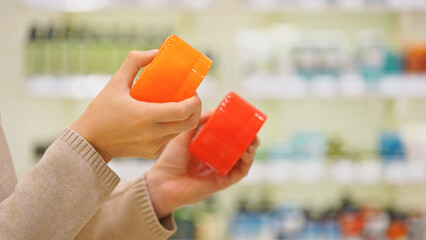 Close up of woman comparing two cosmetic jars in a beauty store, reading labels and ingredients. Female shopper choosing skincare, haircare, or bodycare product. Concept of conscious choice, self-care