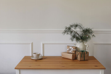 Elegant Christmas table still life. Vase with pine tree branches, gift boxes with ribbons. Cup of coffee, tea. Minimal natural home decoration. Scandinavian style winter decor. Festive interior.