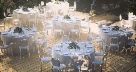 A serene garden venue showcases elegantly arranged tables with white tablecloths and centerpieces, ready for a formal event during the daytime, surrounded by lush greenery.