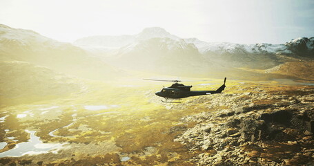 A helicopter glides gracefully over an expansive mountainous terrain bathed in warm sunlight. The rugged landscape features patches of greenery and a serene atmosphere. © icetray