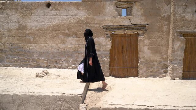 A woman dressed in traditional Arab clothes passes through an archaeological place filled with historical stone structures. The sun shines brightly, illuminating the environment.
