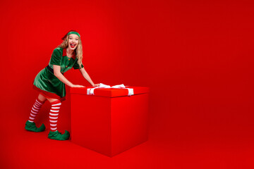 Festive elf poses joyfully with a giant red gift box in a seasonal holiday setting featuring vibrant background and colors