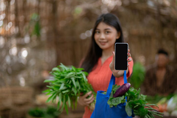 A joyful woman is happily holding fresh vegetables while also using her smartphone and shopping at a vibrant market