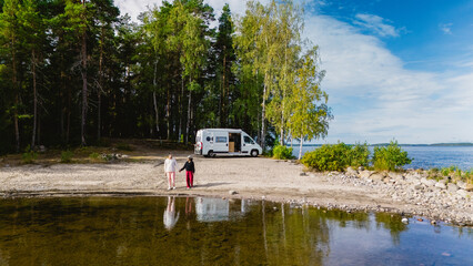 Two friends stroll along the sandy shore of Lake Saimaa, surrounded by lush greenery and a tranquil atmosphere. A camper van is parked nearby, ready for exploration.