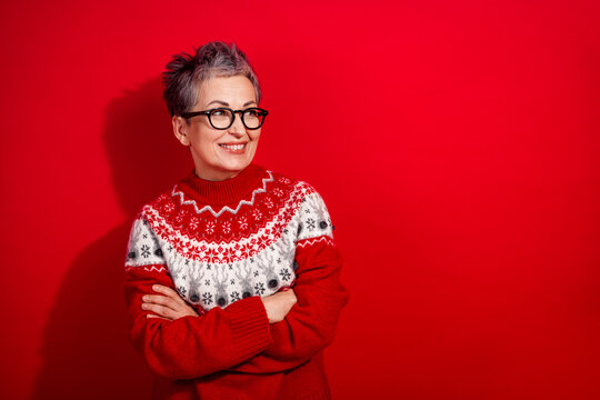 Stylish mature woman in red festive sweater smiling joyfully against bright red background with arms crossed - Powered by Adobe