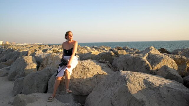 A woman poses on large rocks along the shore as the sun sets over the ocean, creating a tranquil atmosphere with a city skyline visible in the background.