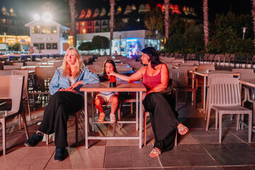 Three females, including a child, sit at an outdoor table at night. One woman reaches toward the child, all lit by colorful event lighting in a relaxed setting.