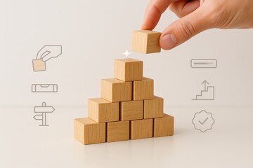 Hand carefully places the final wooden block on top of a pyramid structure made of wooden blocks, symbolizing the completion of a project or reaching a goal against a simple white backdrop.
