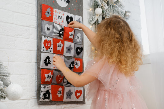 Young girl in pink dress putting gifts in advent calendar pocket, surrounded by festive lights and frosted holiday decor, Christmas celebration 