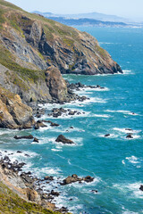 Steep cliffs along the Pacific Ocean near Muir Beach Cove, viewed from Tennessee Valley Trail in Marin Headlands, California. Rugged coastline with waves crashing on rocks under a bright sky
