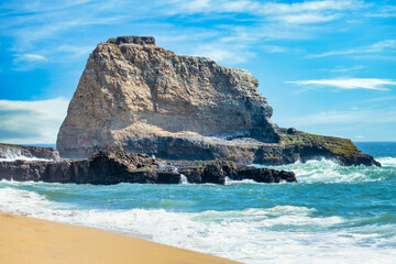 Scenic sandy beach along Highway 1 California with ocean waves crashing on rocks under clear blue sky and coastal cliffs capturing the beauty of the Pacific Coast on a sunny summer day