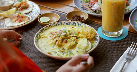 Thai restaurant dining with Khao Soi curry noodles, tacos on wooden table, hand holding spoon. Exotic culinary experience in tropical setting. Cozy, flavorful, Asian cuisine.