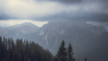 Der graue Himmel verschluckt die Gipfel, ein dichter Nebelschleier verhüllt die Erhabenheit der Berge. Die dunkle Fichtenfront hält stand gegen die feuchte Melancholie des Tals. © Viridi Silva