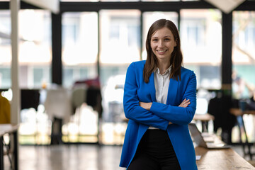 Businesswoman smiling with crossed arms in busy office. Businesswoman is smiling while standing in her company's office.  She is wearing a blue jacket and has her arms crossed.