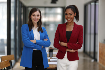 Businesswoman smiling with arms crossed in office. Two confident businesswomen in a modern office, arms crossed, wearing colorful suit, showing teamwork and success.