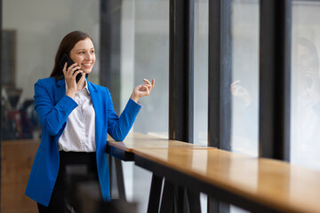 Businesswoman using smartphone and looking through office window. Happy woman taking a break in a modern office, using her smartphone and looking out the window.