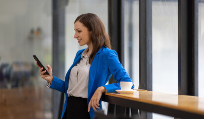 Businesswoman using smartphone and looking through office window. Happy woman taking a break in a modern office, using her smartphone and looking out the window.