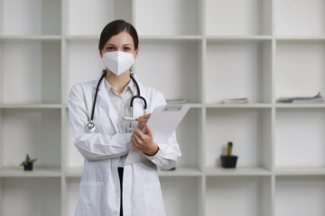 Confident female doctor in a modern clinic, holding a clipboard and wearing a protective mask, conveys reassurance and dedication, providing professional care in a bright and clean office setting.