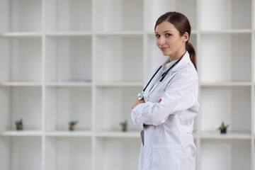 Young female doctor is smiling with her arms crossed in a hospital or doctor's office. She is wearing a stethoscope and a white coat.