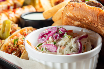 Assorted fast food on a table: tacos with toppings, burger with fries, creamy coleslaw in ramekin, and dipping sauce. Casual dining concept, vibrant and appetizing