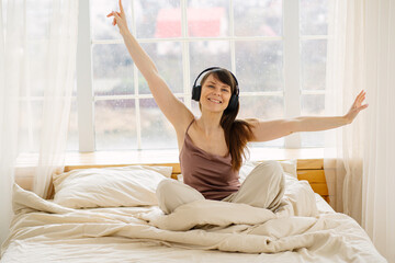 Beautiful young woman relaxing in bed with headphones, enjoying music after waking up. Cozy morning...