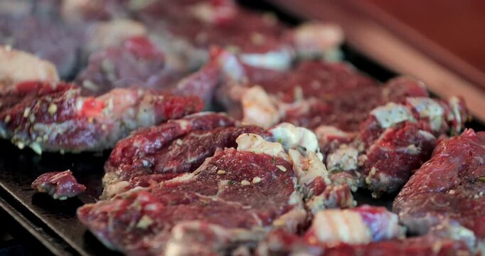 Various cuts of raw beef marinated and laid out on grill for cooking, showing spices, fat, and red meat texture in high-resolution close-up frame
